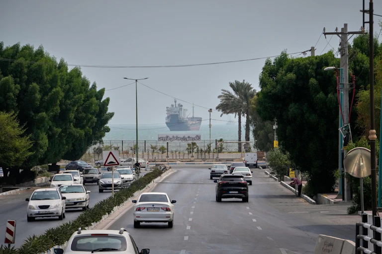 Cars on a road heading towards thye Strait of Hormuz where a tanker sits moored.