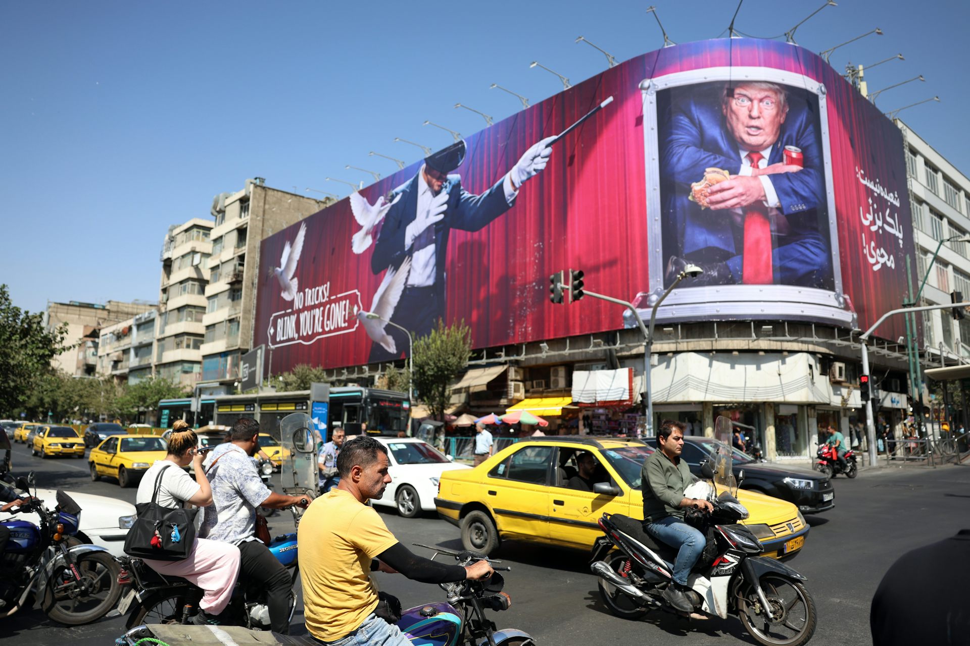 People drives cars and motorcycles in the centre of Iran past an anti-Trump banner.