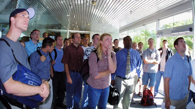 Several people in a hall look up as they watch television.