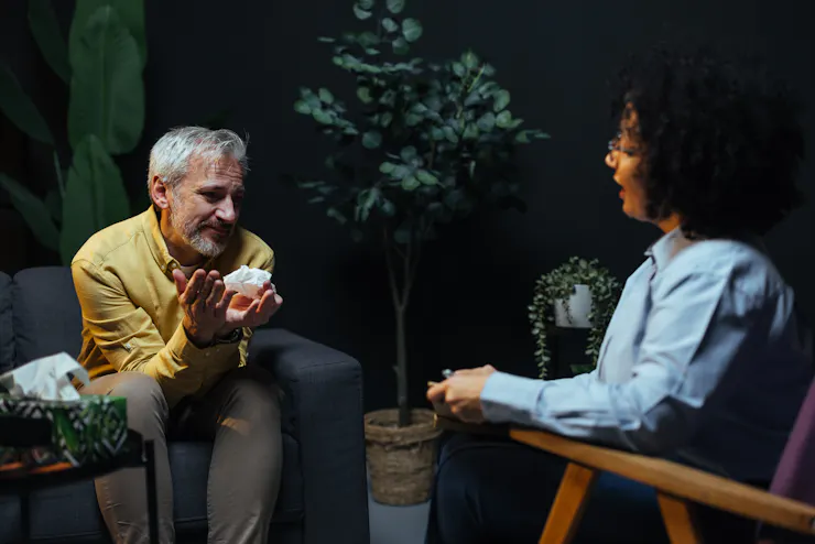 Older man sitting in an armchair across from a therapist, talking and holding a tissue