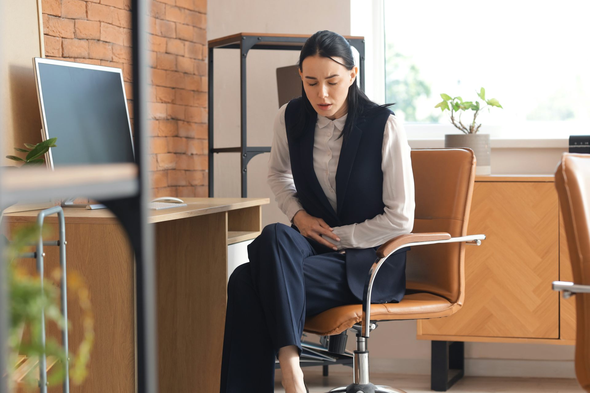 Jeune femme, assise à son bureau dans un cadre professionnel, se tenant le ventre de douleur.