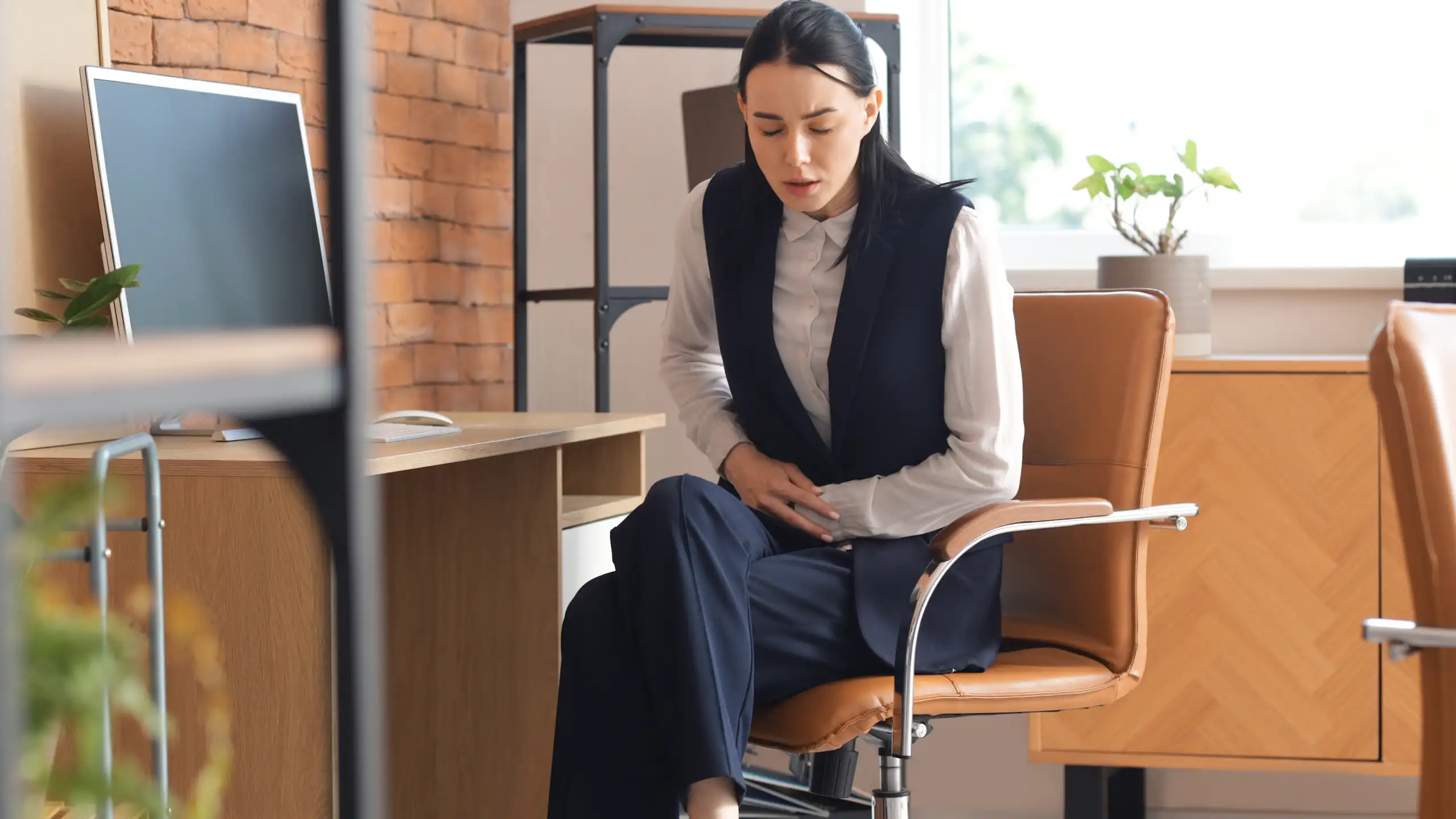 Jeune femme, assise à son bureau dans un cadre professionnel, se tenant le ventre de douleur.