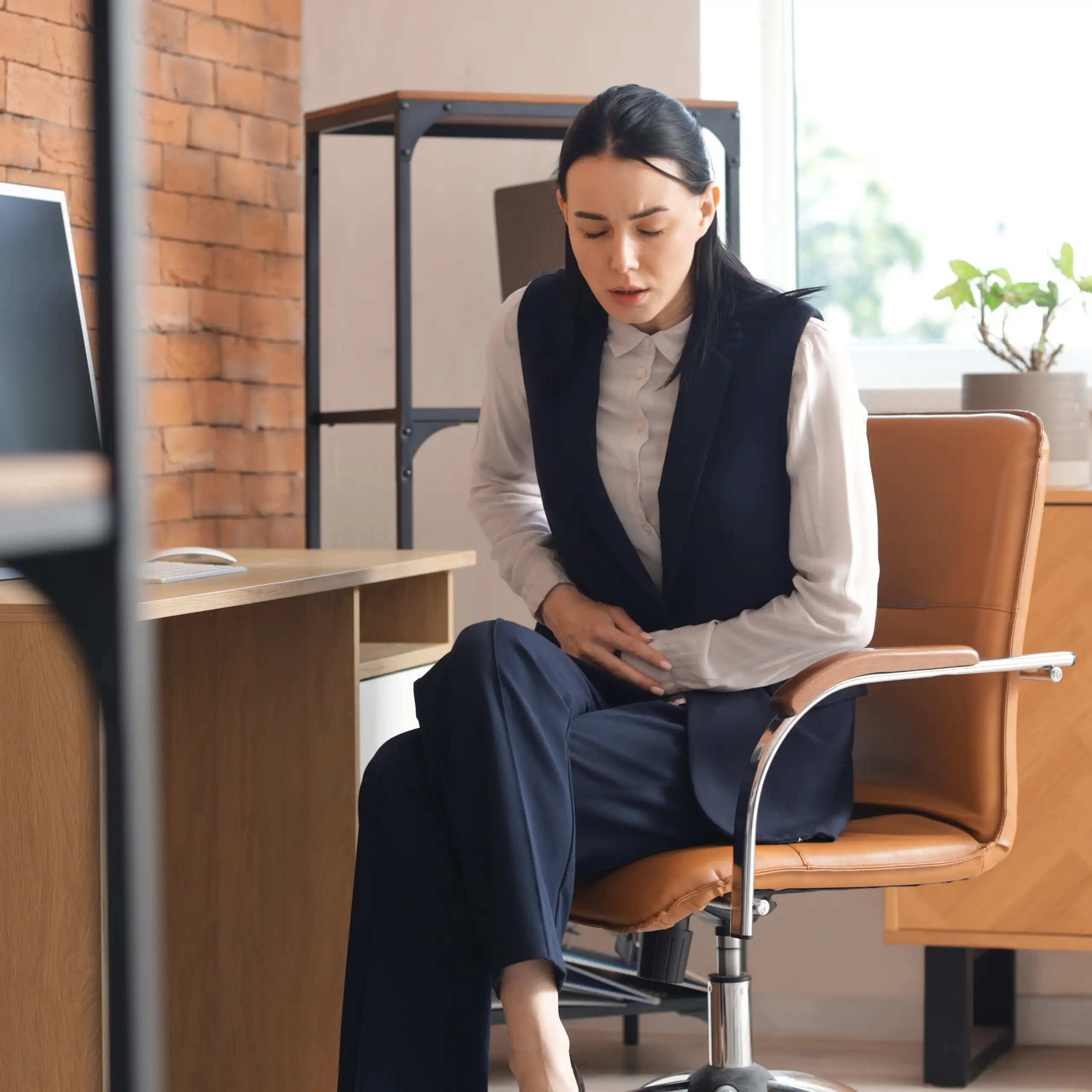Jeune femme, assise à son bureau dans un cadre professionnel, se tenant le ventre de douleur.