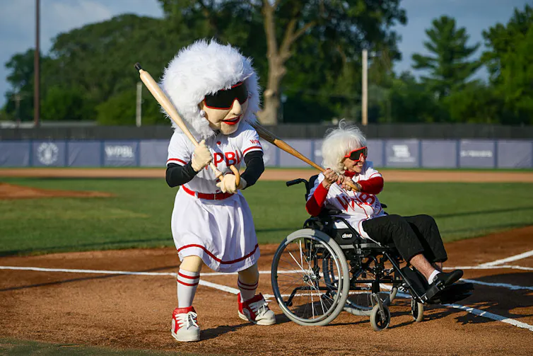 Large white-haired mascot with huge sunglasses poses with a bat next to a woman in a wheelchair also wearing the same sunglasses and posing with a bat.