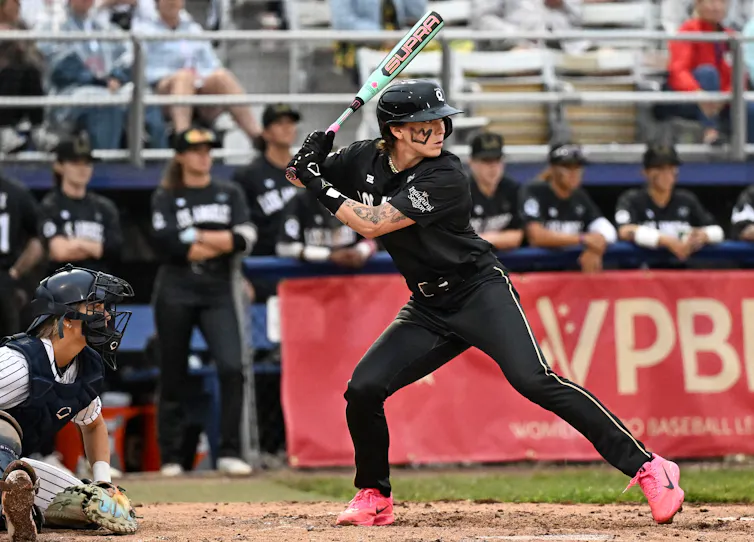 Young woman wearing black uniform anticipates a pitch while holding a baseball bat.