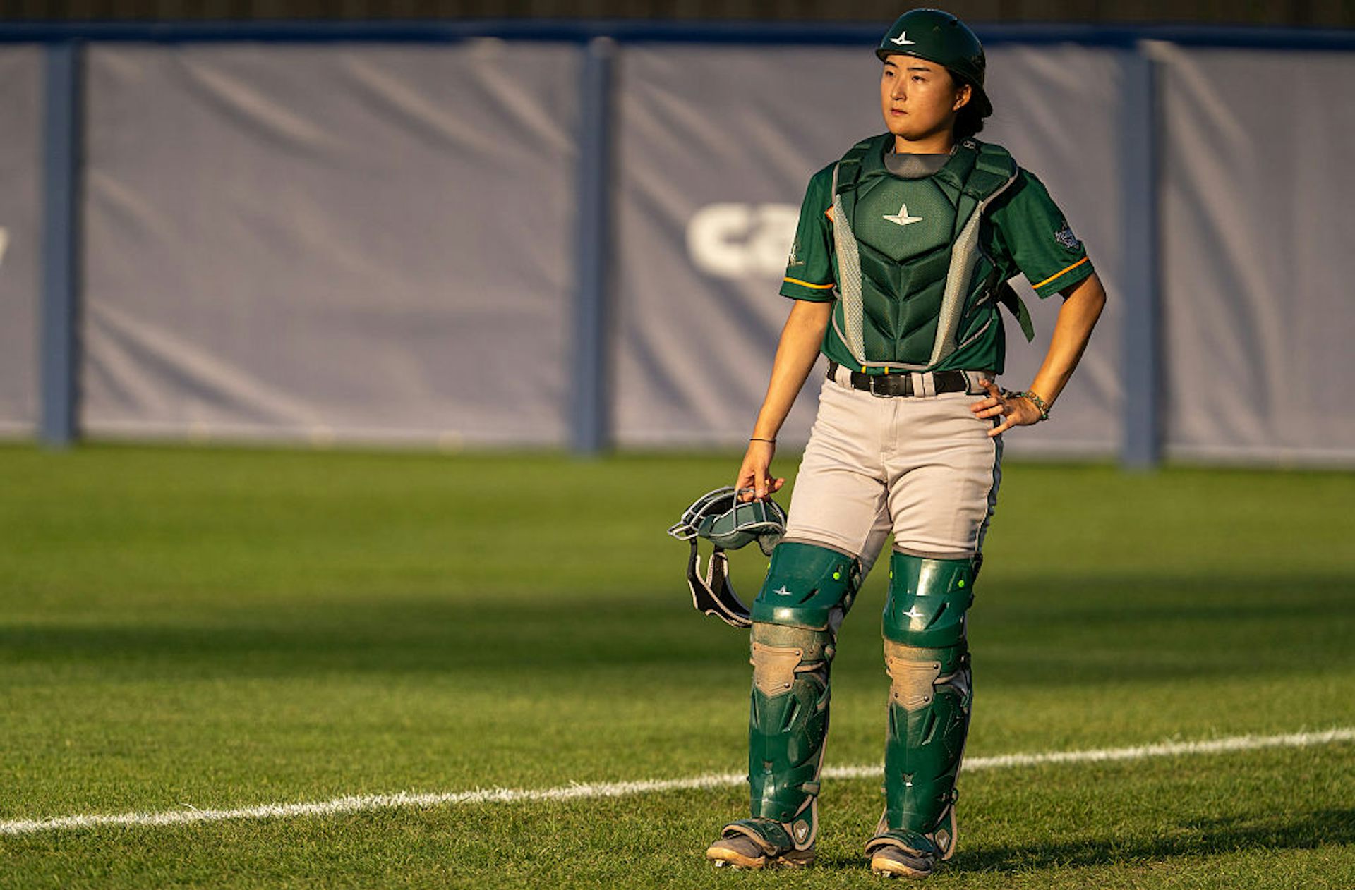 Young Asian woman wearing green catching gear stands on a baseball field.
