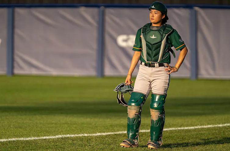 Young Asian woman wearing green catching gear stands on a baseball field.