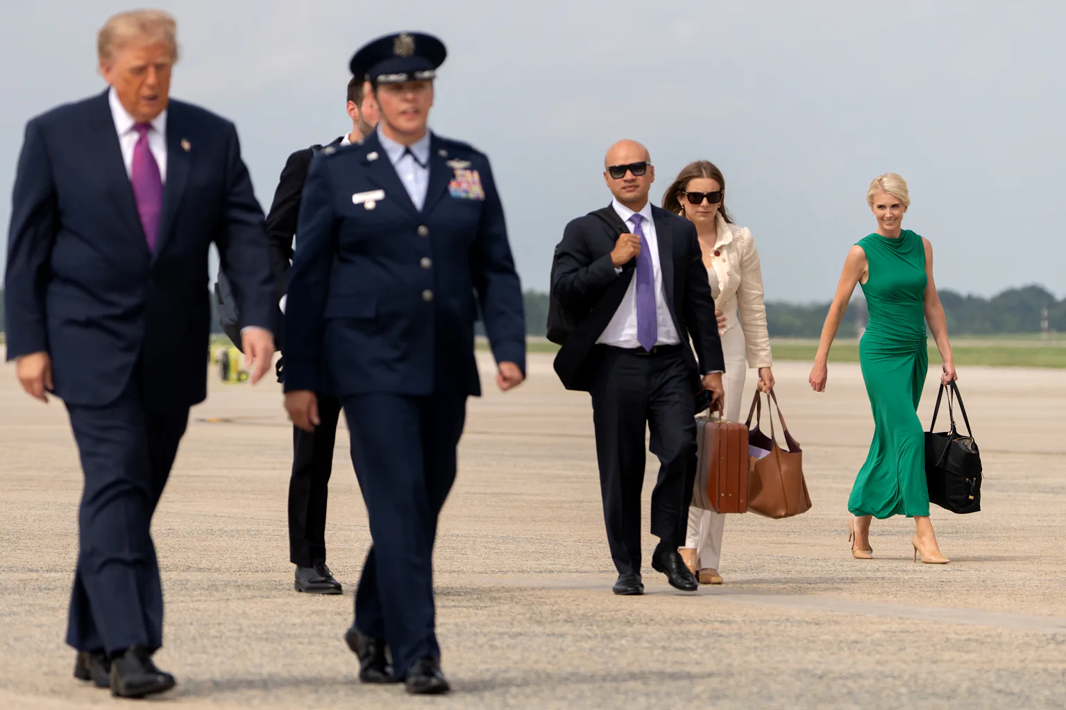 President Donald Trump walks across a tarmac, followed by several others, including Natalie Harp at right.