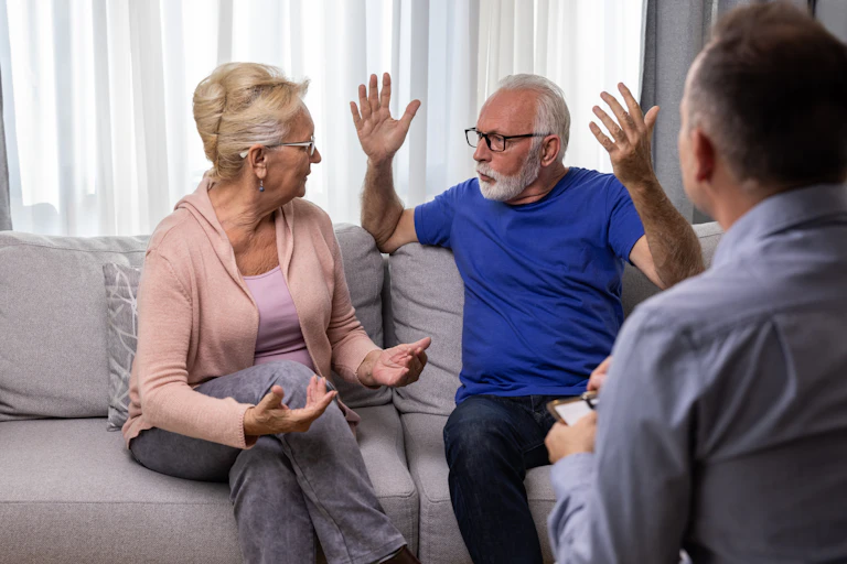 An older couple, woman and man, sit arguing on a therapists's couch during their session.