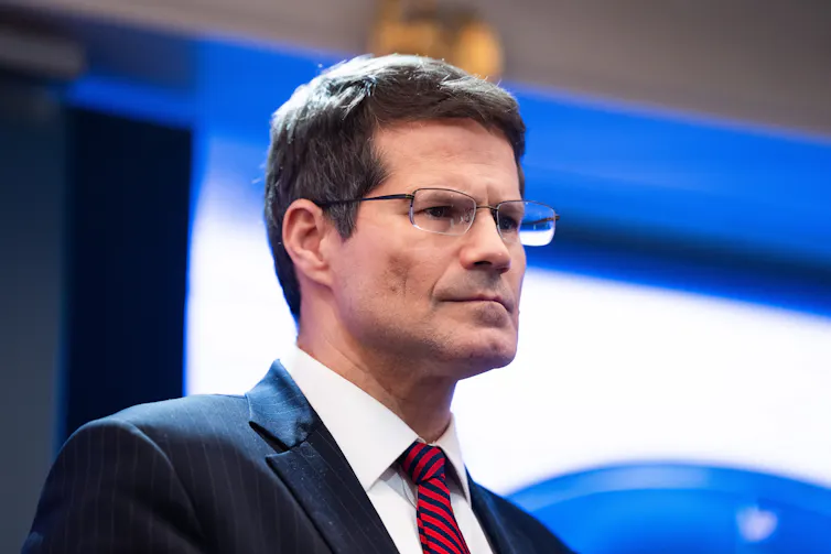 A serious-looking man with dark hair and glasses, wearing a blue jacket, white shirt and red tie.