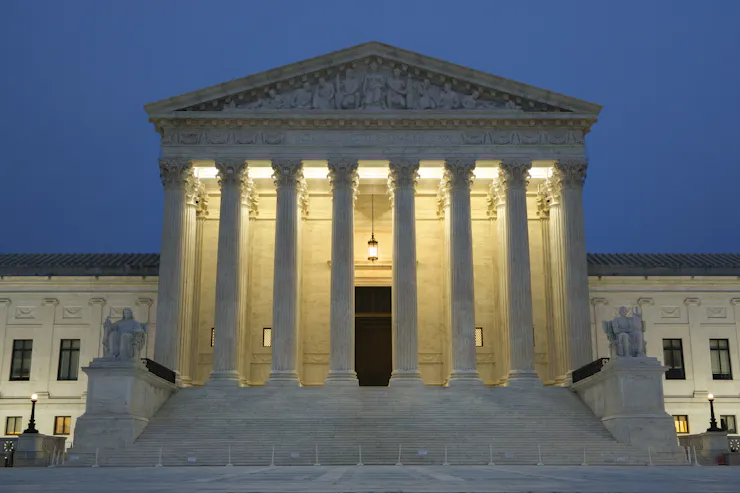 The Supreme Court building is seen at dusk, with lights illuminating behind its pillars.