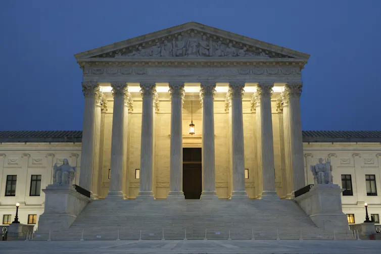The Supreme Court building is seen at dusk, with lights illuminating behind its pillars.