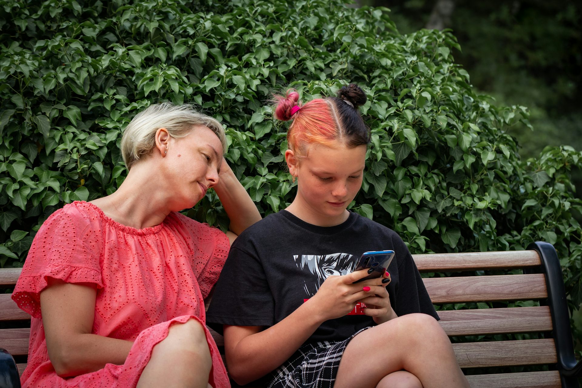 A mother watches her teen scroll their phone on a park bench