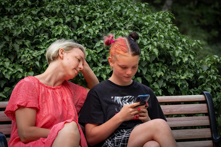 A mother watches her teen scroll their phone on a park bench