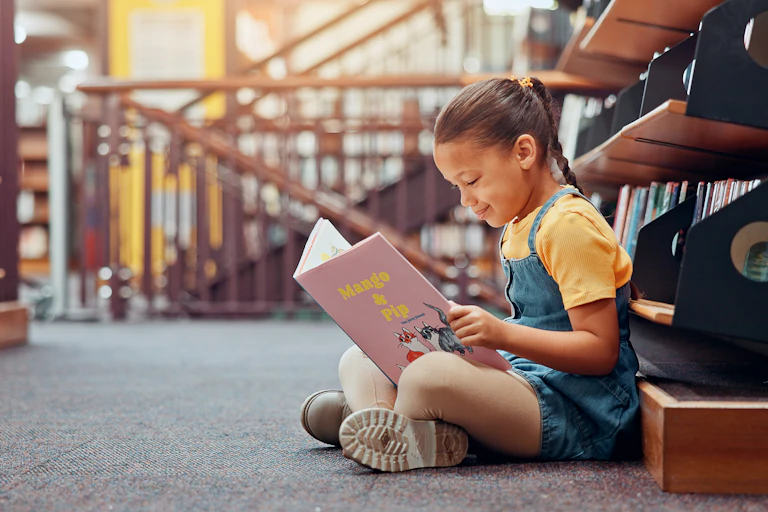 Una nena llig un llibre asseguda a terra.
