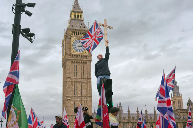 A protestor holds up a Christian cross and a union jack against the sky in front of Big Ben at the Unite the Kingdom 2026 rally in Parliament Square, London