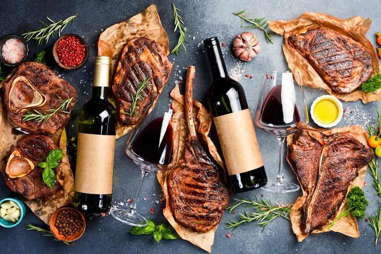 Top view of bottles of red wine and aged beef steaks surrounded by cloves of garlic and sprigs of rosemary on a grey stone background.