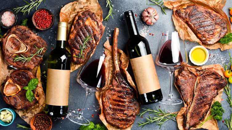 Top view of bottles of red wine and aged beef steaks surrounded by cloves of garlic and sprigs of rosemary on a grey stone background.