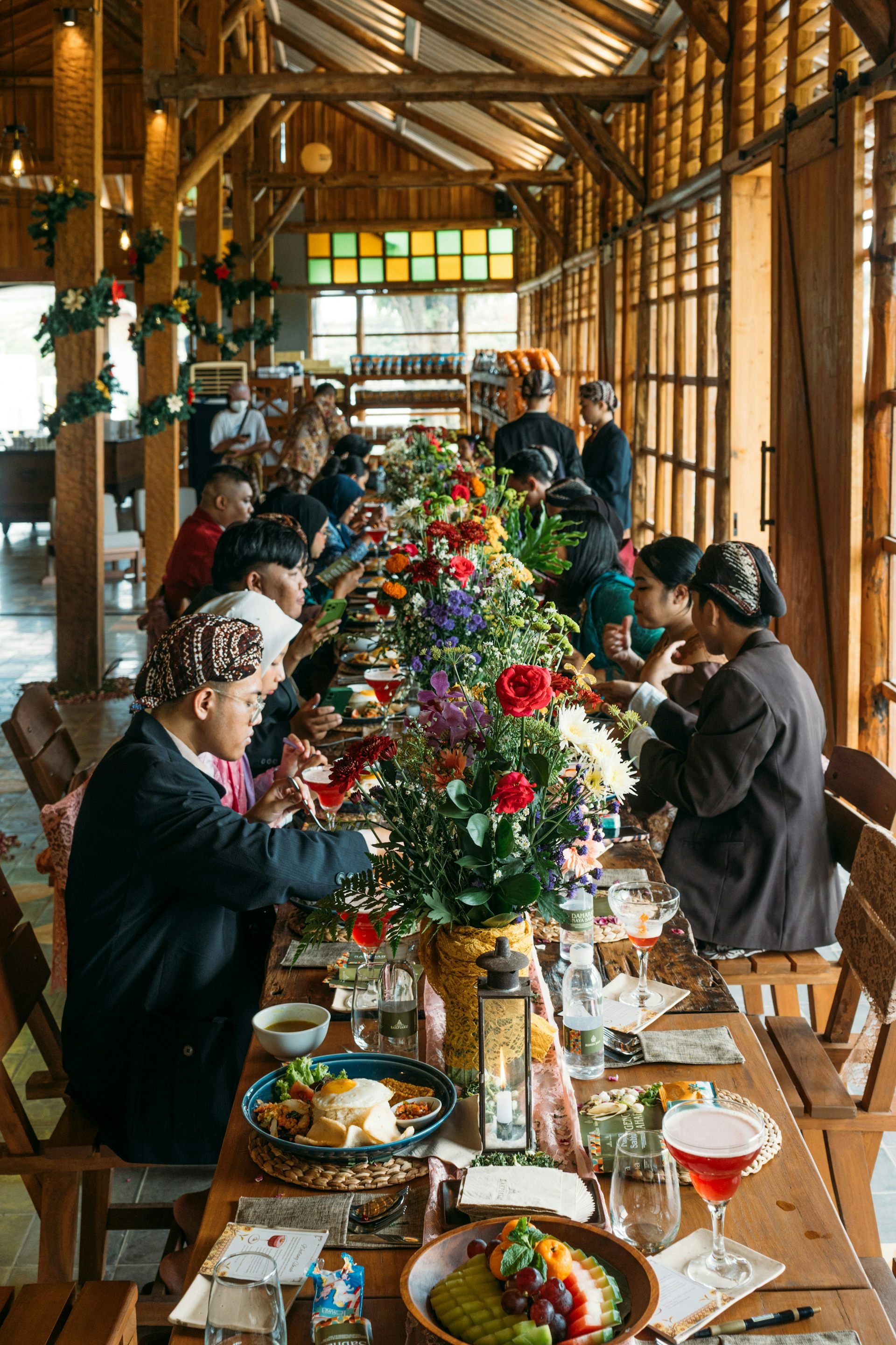 At a long table covered by flowers and plates of food, a diverse group of travelers shares a meal.