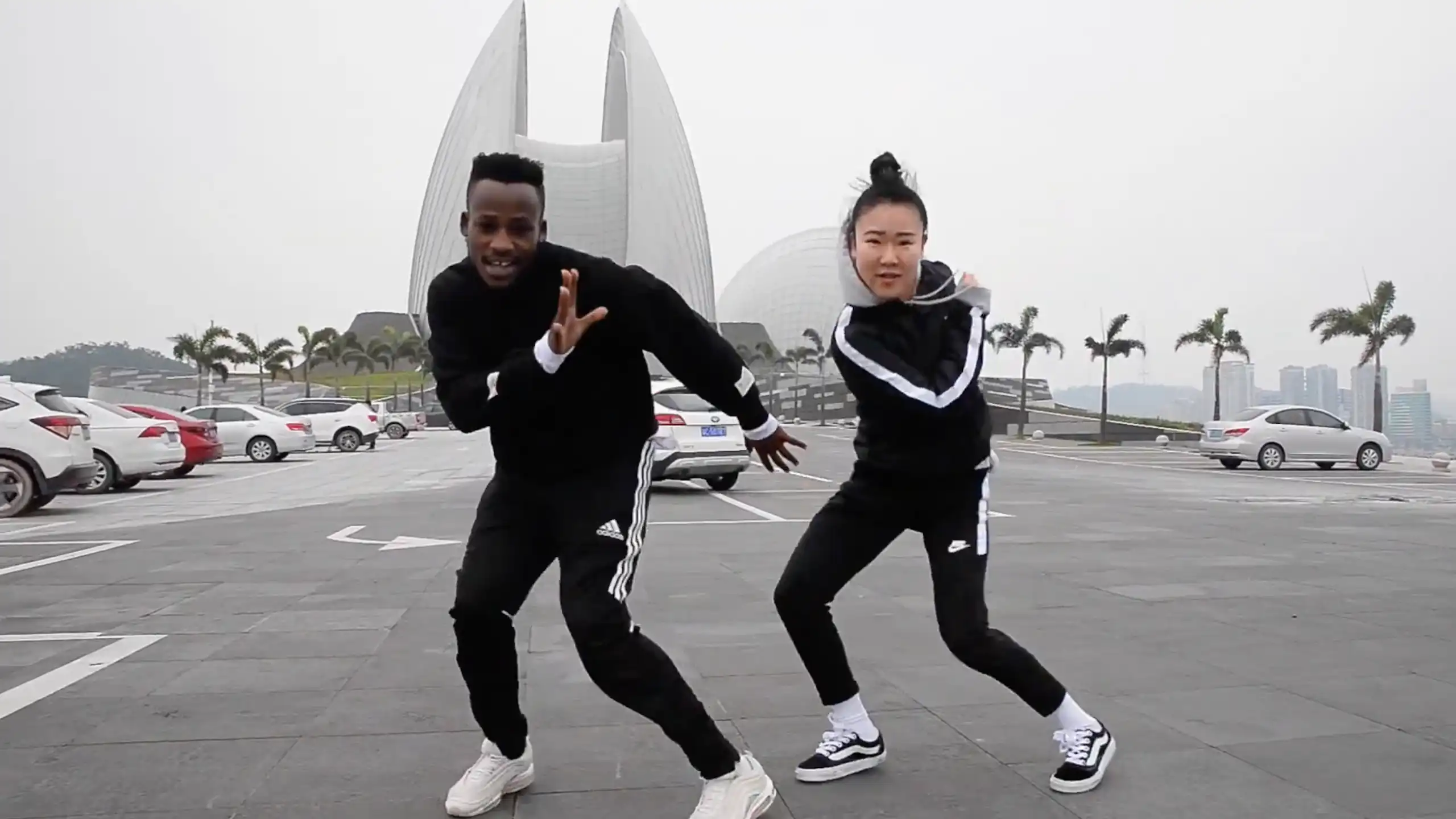 An African man and a Chinese women dressed in black tracksuits dance in front of a futuristic building.
