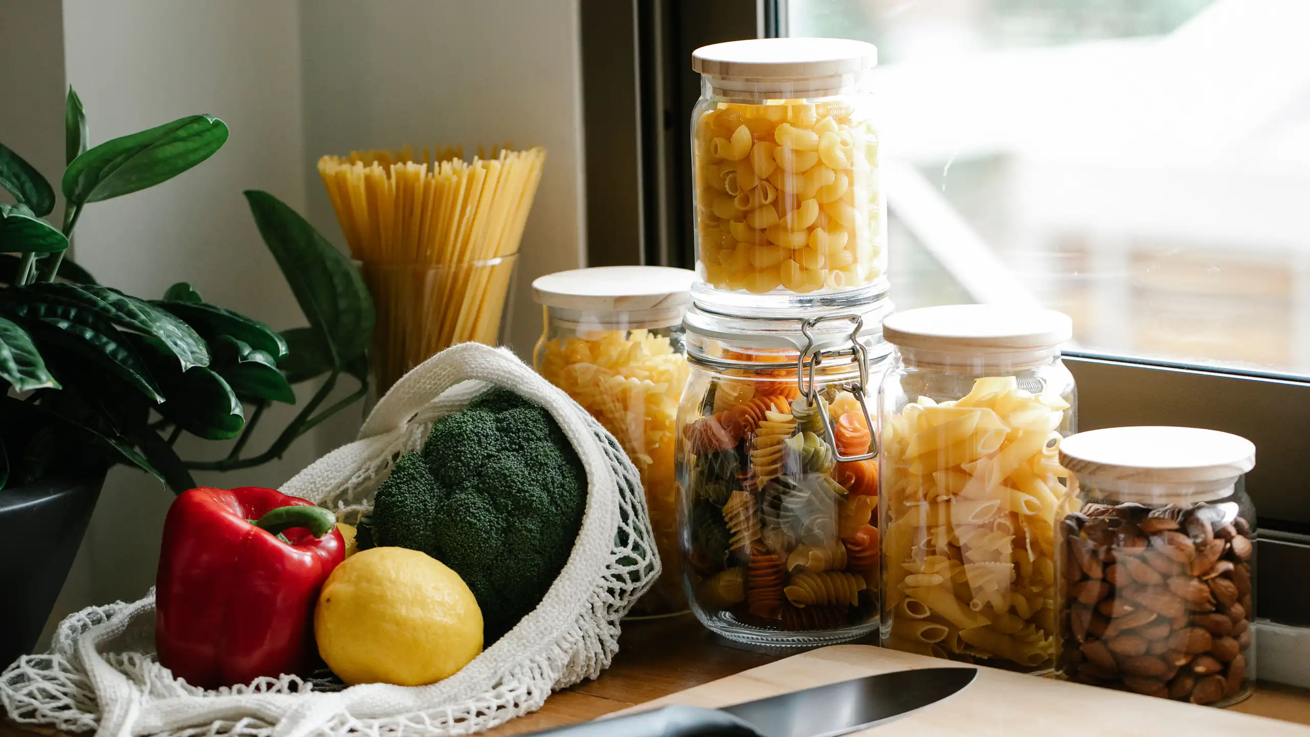 Diferentes tipos de massas em potes de vidro e um saco com legumes sobre um armário de cozinha, ao lado de uma tábua de corte e uma faca, em frente a uma janela