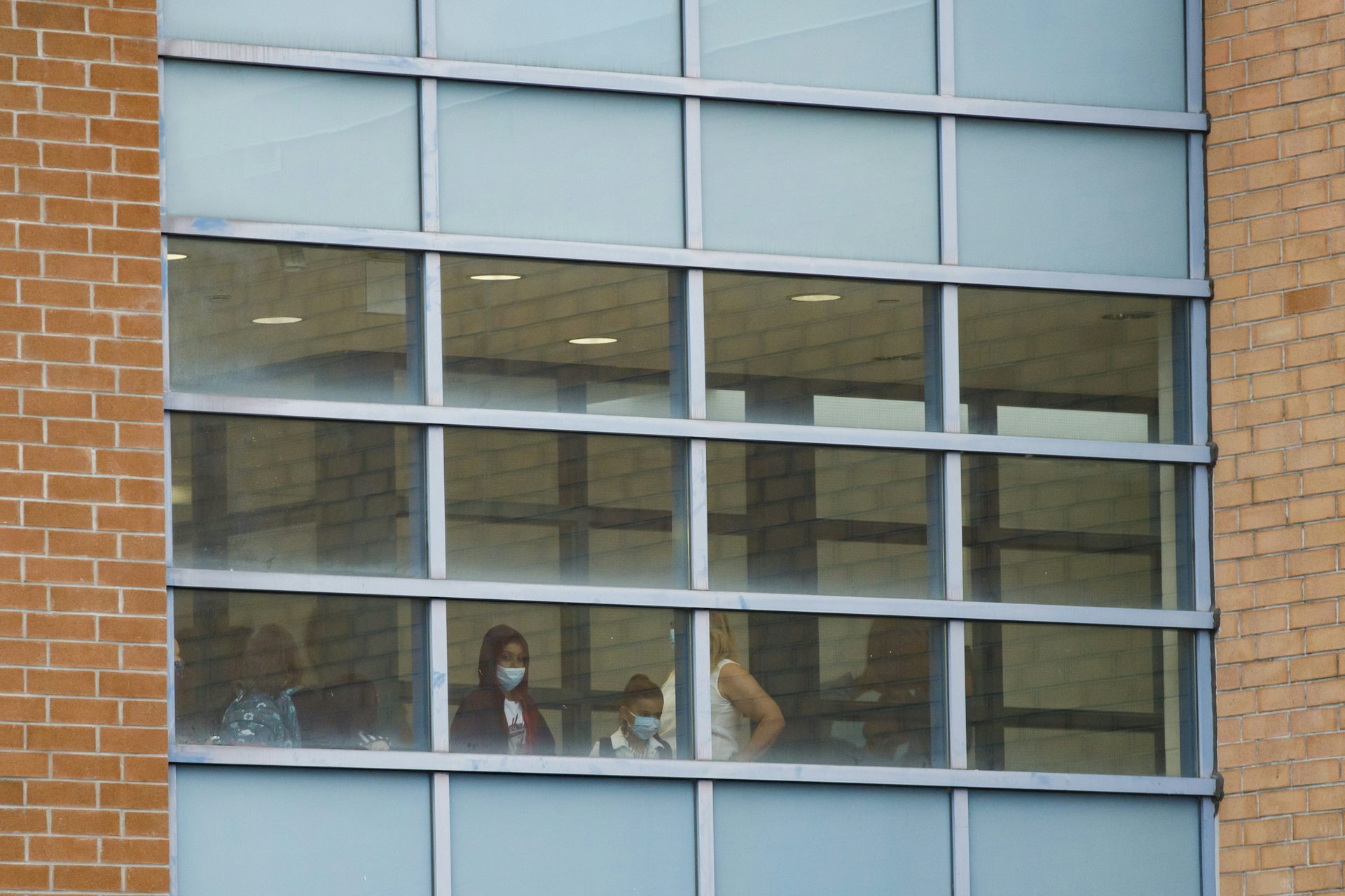 Students and an educator in a facemask look out the window of a school.