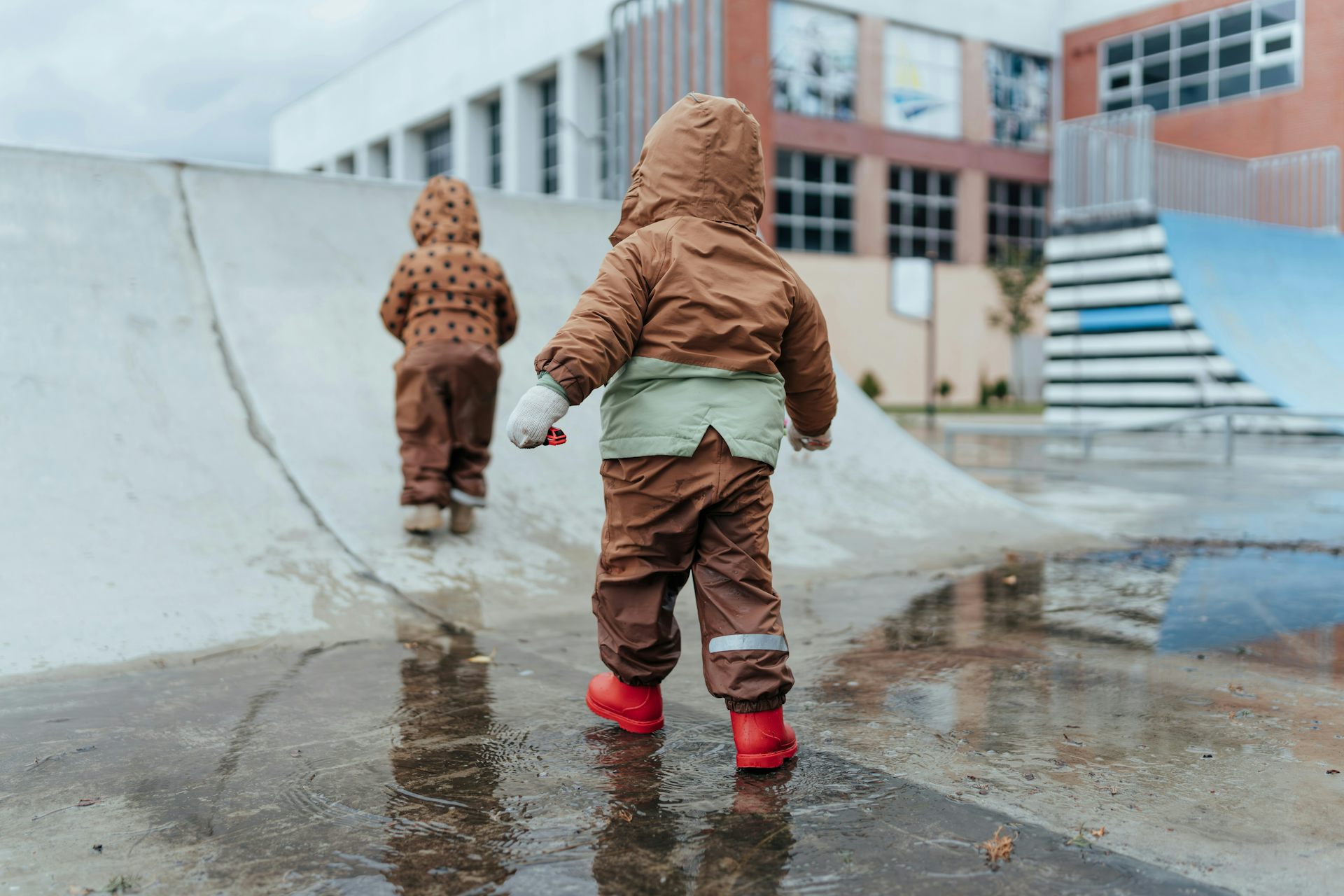 Two children in all-weather suits seen on a skateboard ramp next to a large building.