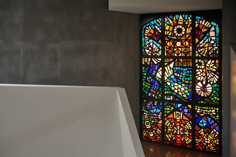 Interior shot of a stained-glass window with nine panels, installed in a grey concrete gallery wall.