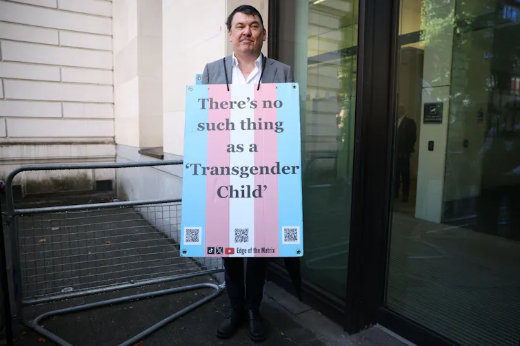 a man in a shirt and suit jacket, by a trolley, wearing a sandwich board that reads: 'There's no such thing as a transgender child.'