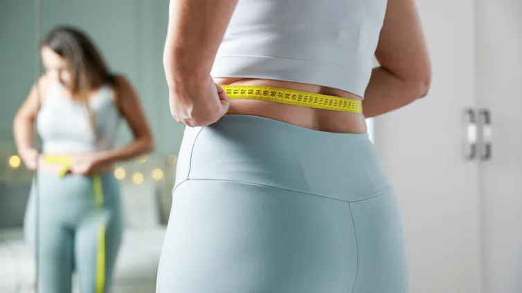Woman in exercise Lycra standing before mirror, tape measure around waist