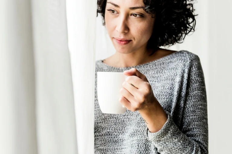 A woman holding a white mug looking past a curtain
