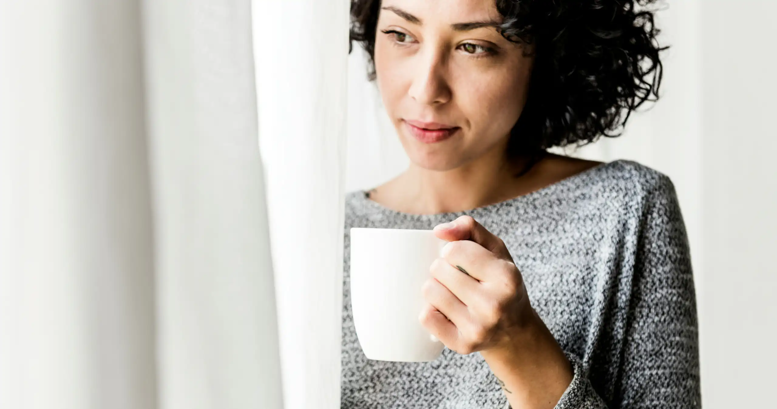 A woman holding a white mug looking past a curtain