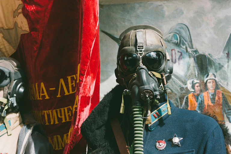 A man dressed in army fatigues and wearing a gas mask while standing in front of a red flag and a painting of people coming out of a plane