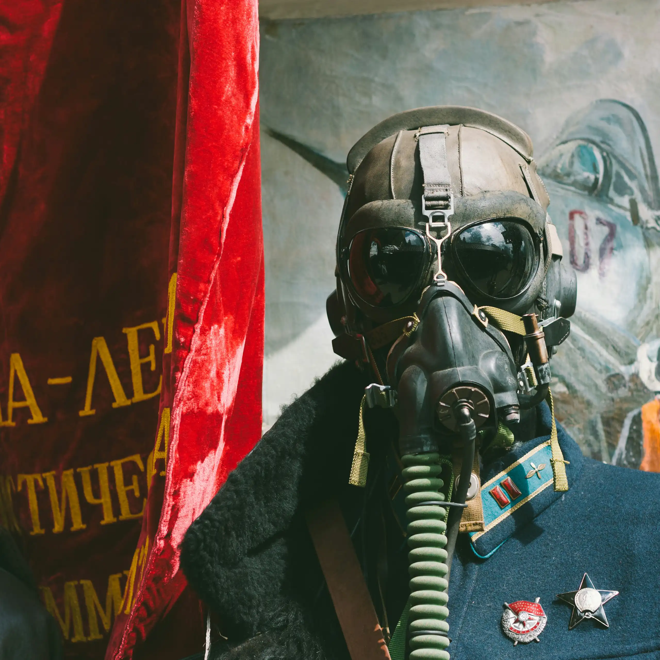 A man dressed in army fatigues and wearing a gas mask while standing in front of a red flag and a painting of people coming out of a plane