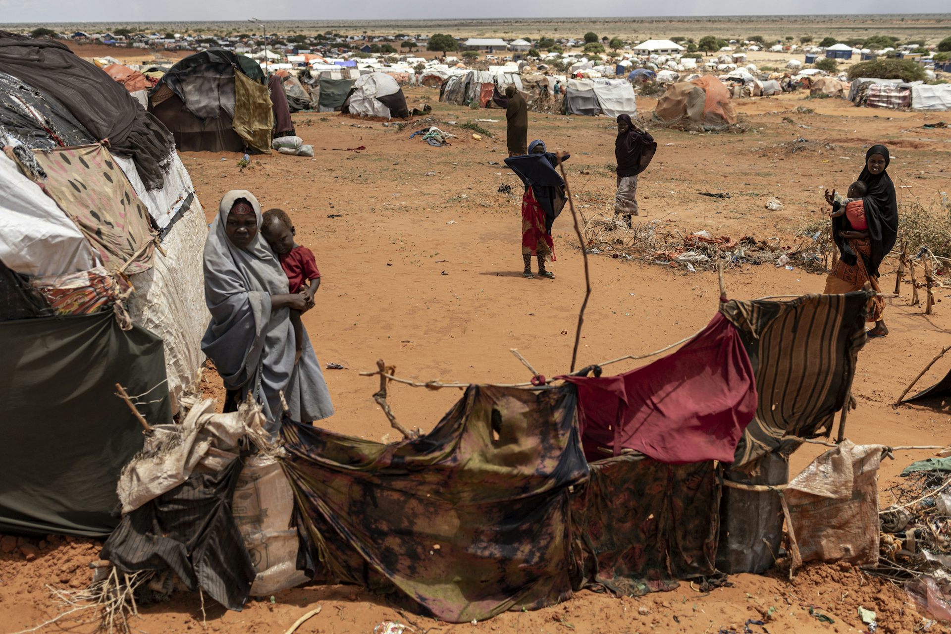 Dry, treeless landscape with small shelters made out of cloths and sticks. A few women standing among them.