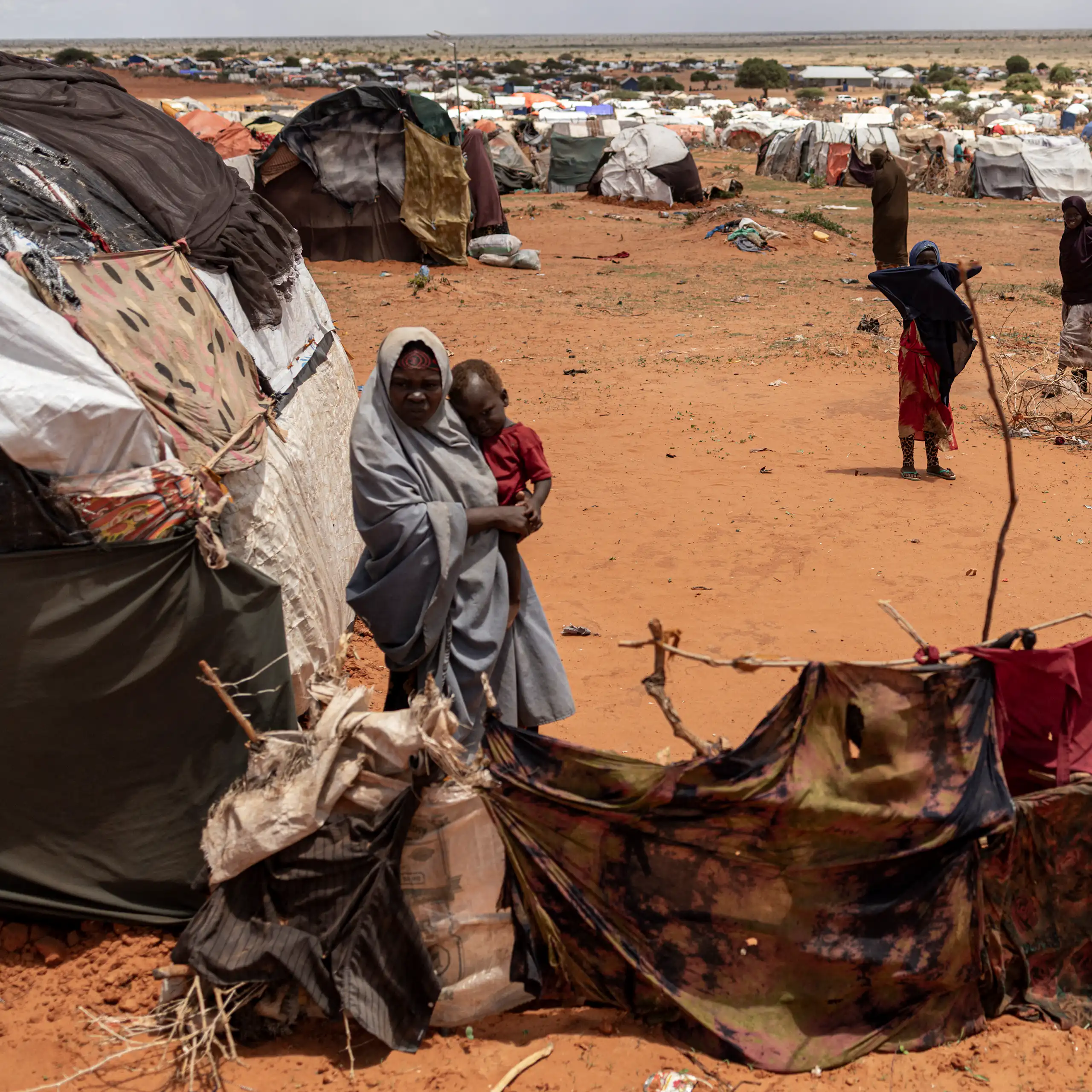 Dry, treeless landscape with small shelters made out of cloths and sticks. A few women standing among them.