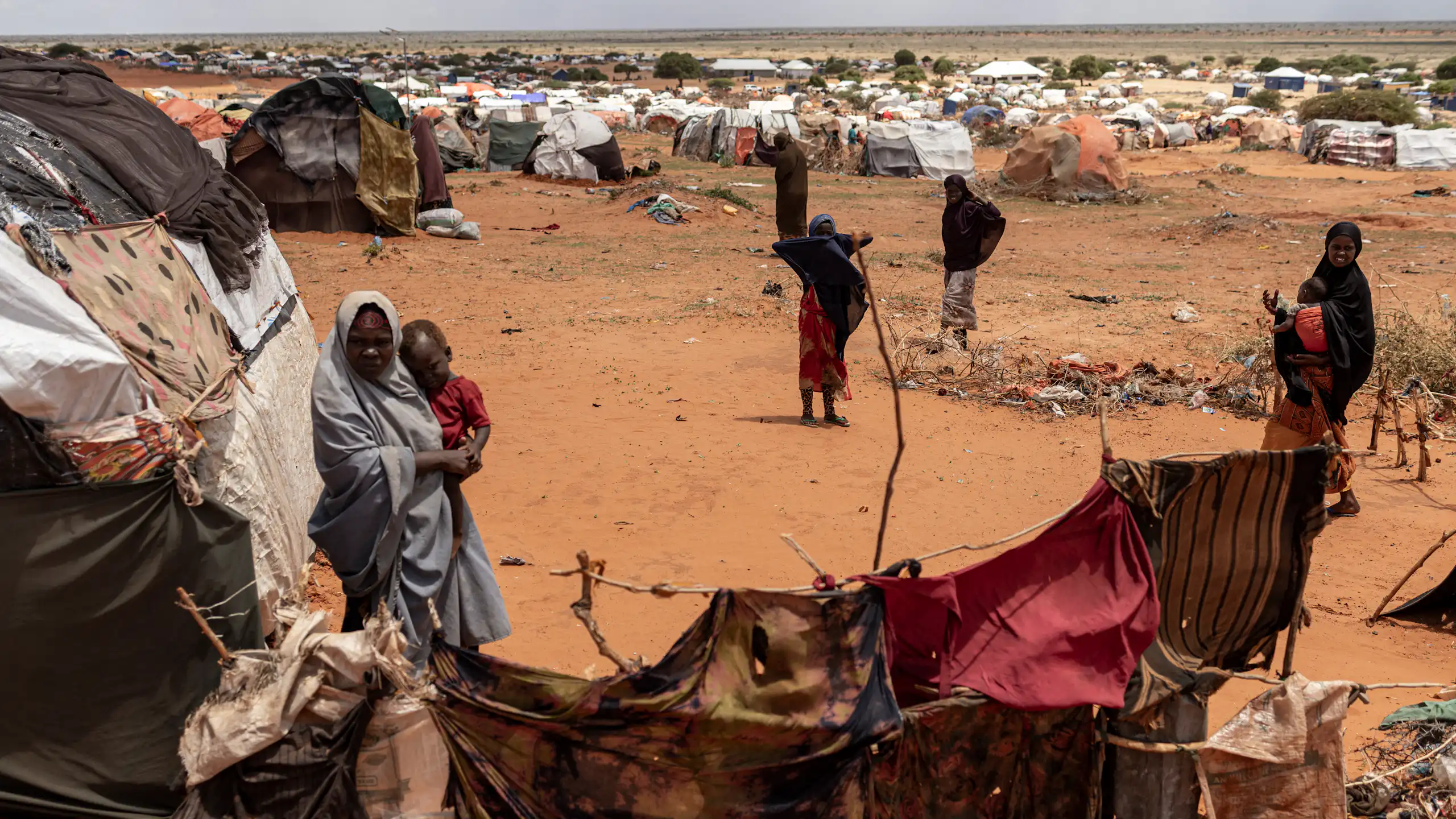 Dry, treeless landscape with small shelters made out of cloths and sticks. A few women standing among them.