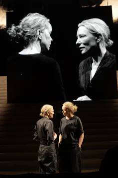 Two women on stage staring at each other, the two projected on screen behind themselves.