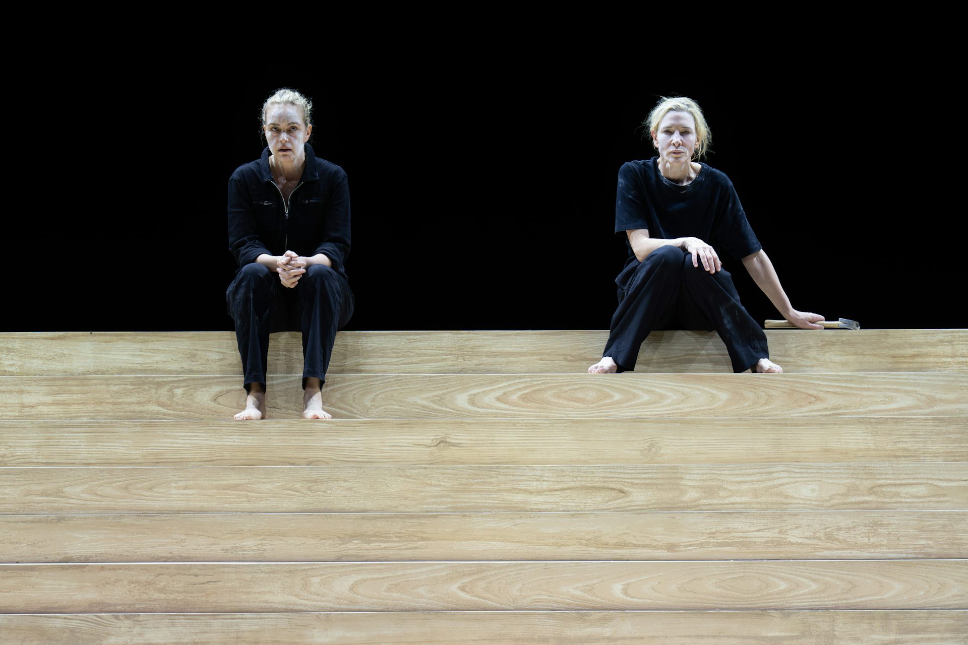 Two women sit on some wooden steps on stage against a black backdrop.