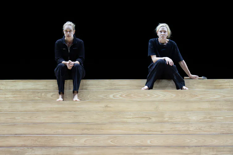 Two women sit on some wooden steps on stage against a black backdrop.