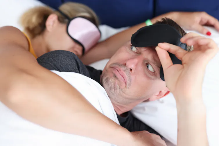man in bed lifts eye mask and looks quizzically over at woman asleep in eye mask