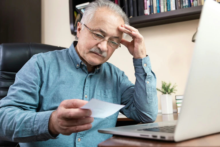 An older man looks at a paper statement, with a confused expression, in front of his laptop.