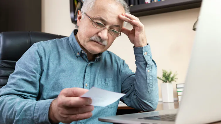 An older man looks at a paper statement, with a confused expression, in front of his laptop.