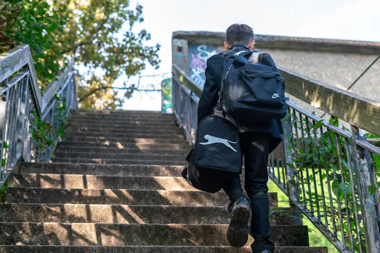 schoolboy seen from behind climbing stairs to a footbridge carrying a rucksack and a sports bag.