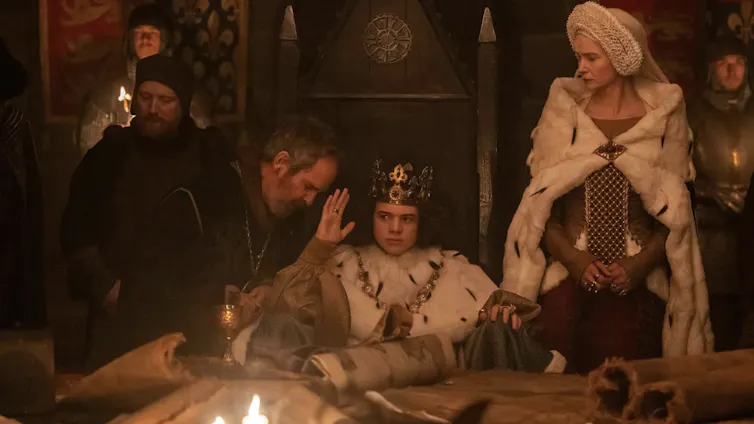 Movie still of a young king who's seated and wearing a crown and robe, surrounded by his courtiers in a dimly lit room.