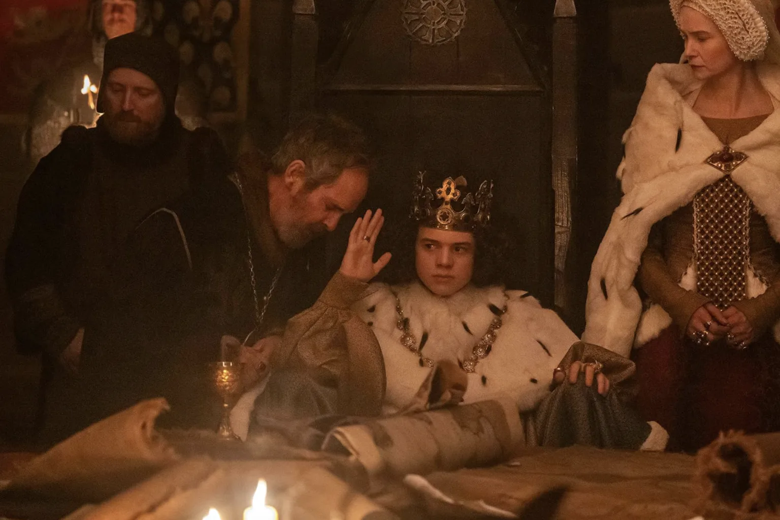 Movie still of a young king who's seated and wearing a crown and robe, surrounded by his courtiers in a dimly lit room.