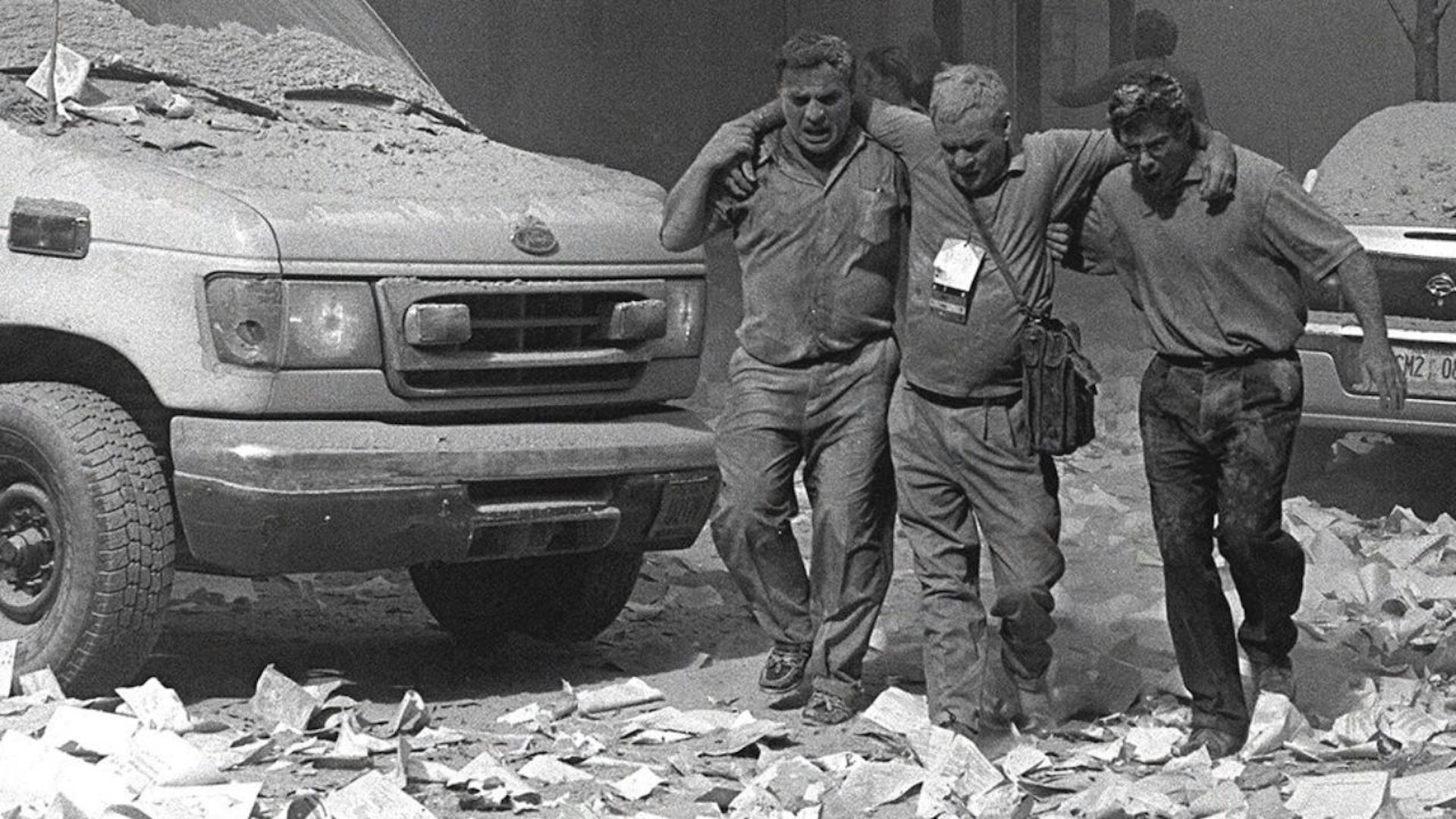 Two men are helping a third  man who is injured. They are walking through debris past a truck. They are covered in ash falling from the September 11 attacks. The photo is black and white.