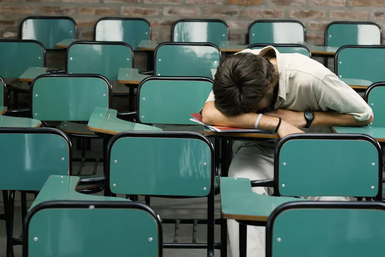 A person rests their head on their arms at a desk in a classroom full of empty teal desks and connected chairs.