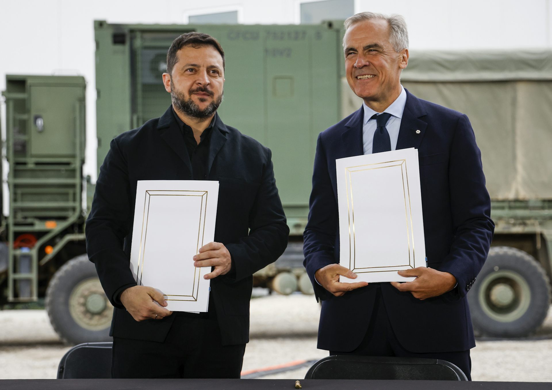 Two white men, one middle-aged and one older with grey hair, stand smiling while holding up documents