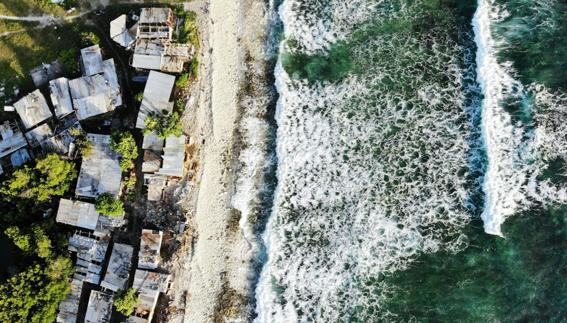 Aerial view of homes in Tuvulu, next to the Pacific Ocean, with ocean waves crashing on the right.